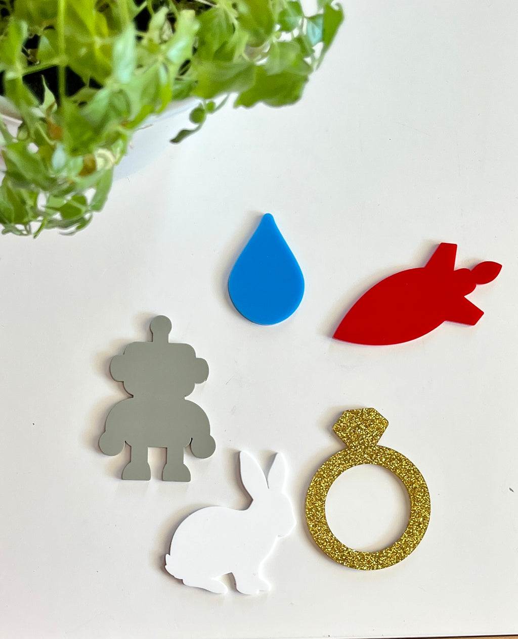 Glossy acrylic shapes featuring gray robot, blue drop, red rocket, white rabbit, and gold glitter ring on white surface with green plant above
