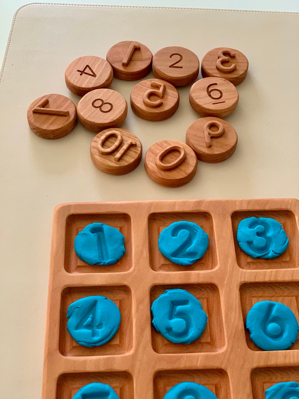 Wooden number discs arranged above a wooden tray holding blue clay imprints of numbers 1 through 6 on a beige surface