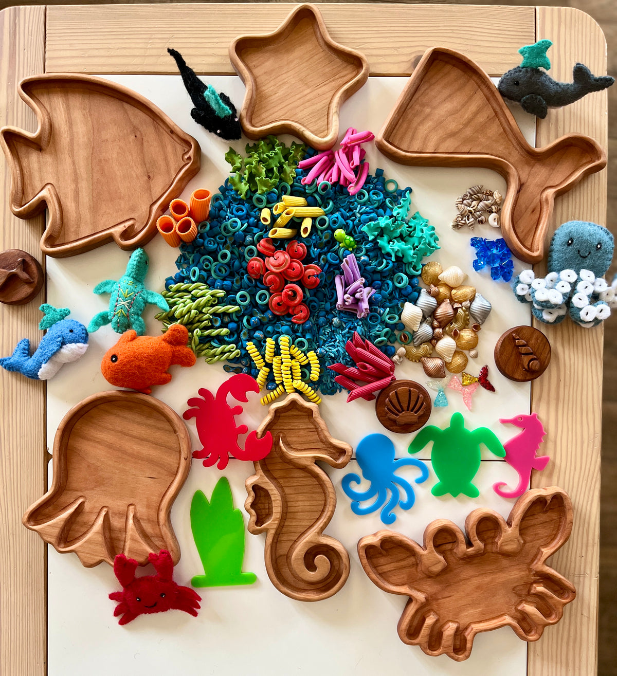 Wooden sea animal-shaped trays with colorful pasta, shells, and felt sea creature toys arranged on a board