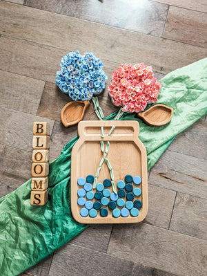 Natural wooden 1.5-inch alphabet blocks stacked to spell BLOOMS with decorative wooden flowers and jar on wooden floor background