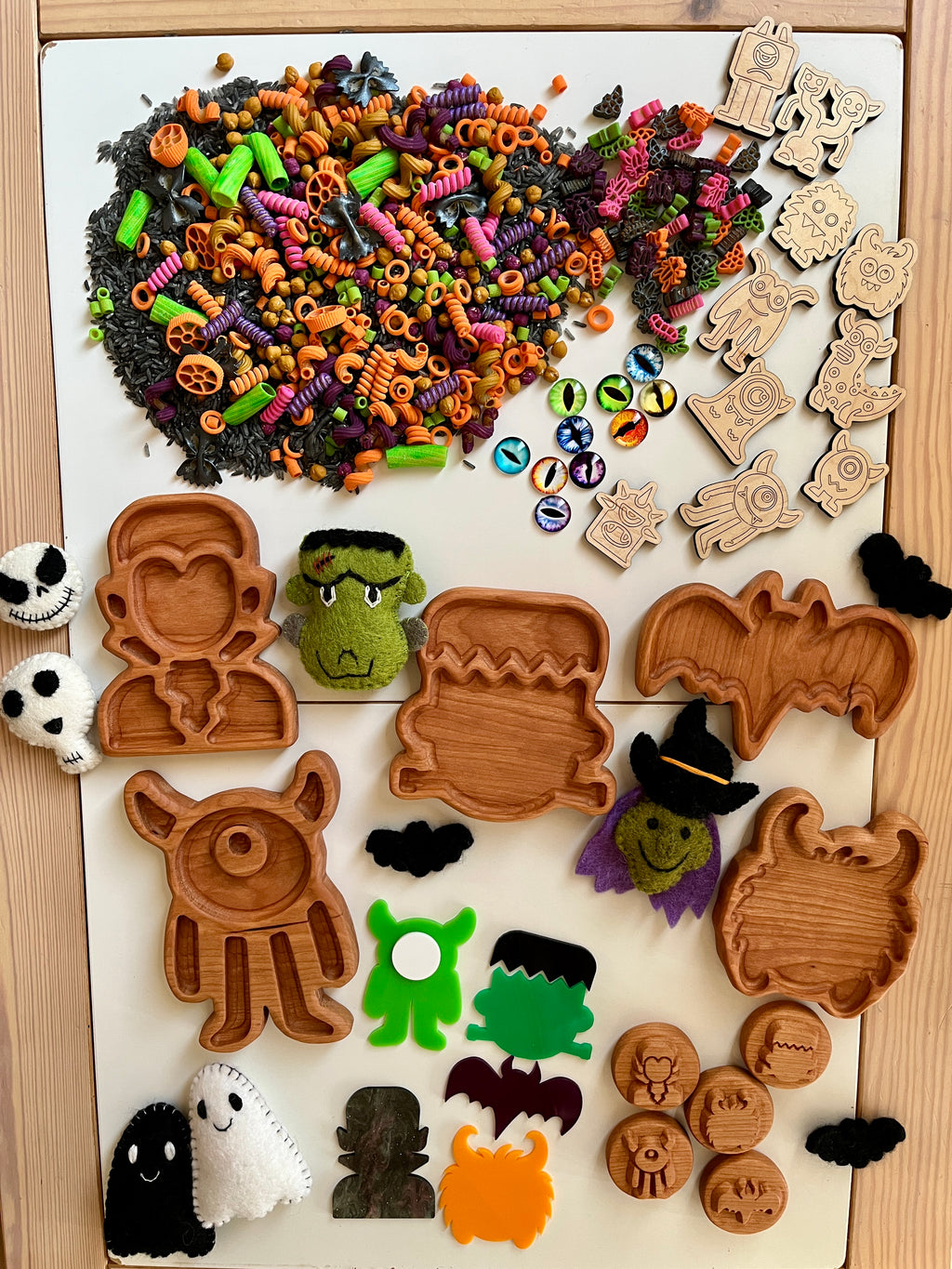 Halloween-themed wooden trays, colorful pasta shapes, felt ghosts, monsters, and decorative craft stamps on wooden table.