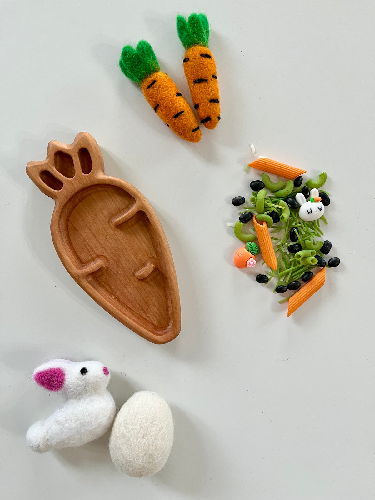 Wooden carrot-shaped tray surrounded by felt carrots, a felt bunny, an egg, and small pasta and bean mix on a white surface