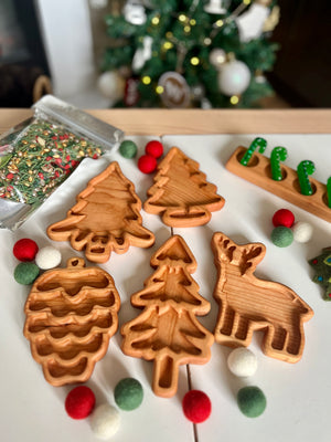 Wooden baking molds shaped like pinecone, Christmas trees, and a reindeer on white surface with holiday decor nearby
