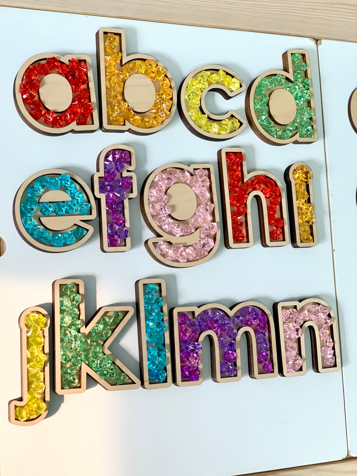 Laser cut wooden alphabet sensory trays filled with colorful translucent beads on white board background