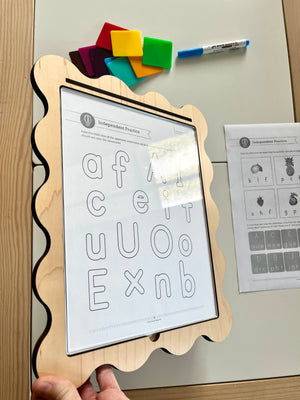 Wooden scalloped-edge frame holding clear sheet over alphabet practice worksheet with colorful magnetic tiles and marker nearby