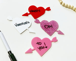 Four arrow-pierced heart-shaped boards in red, white, pink, and glitter pink with a handwritten Valentine’s message