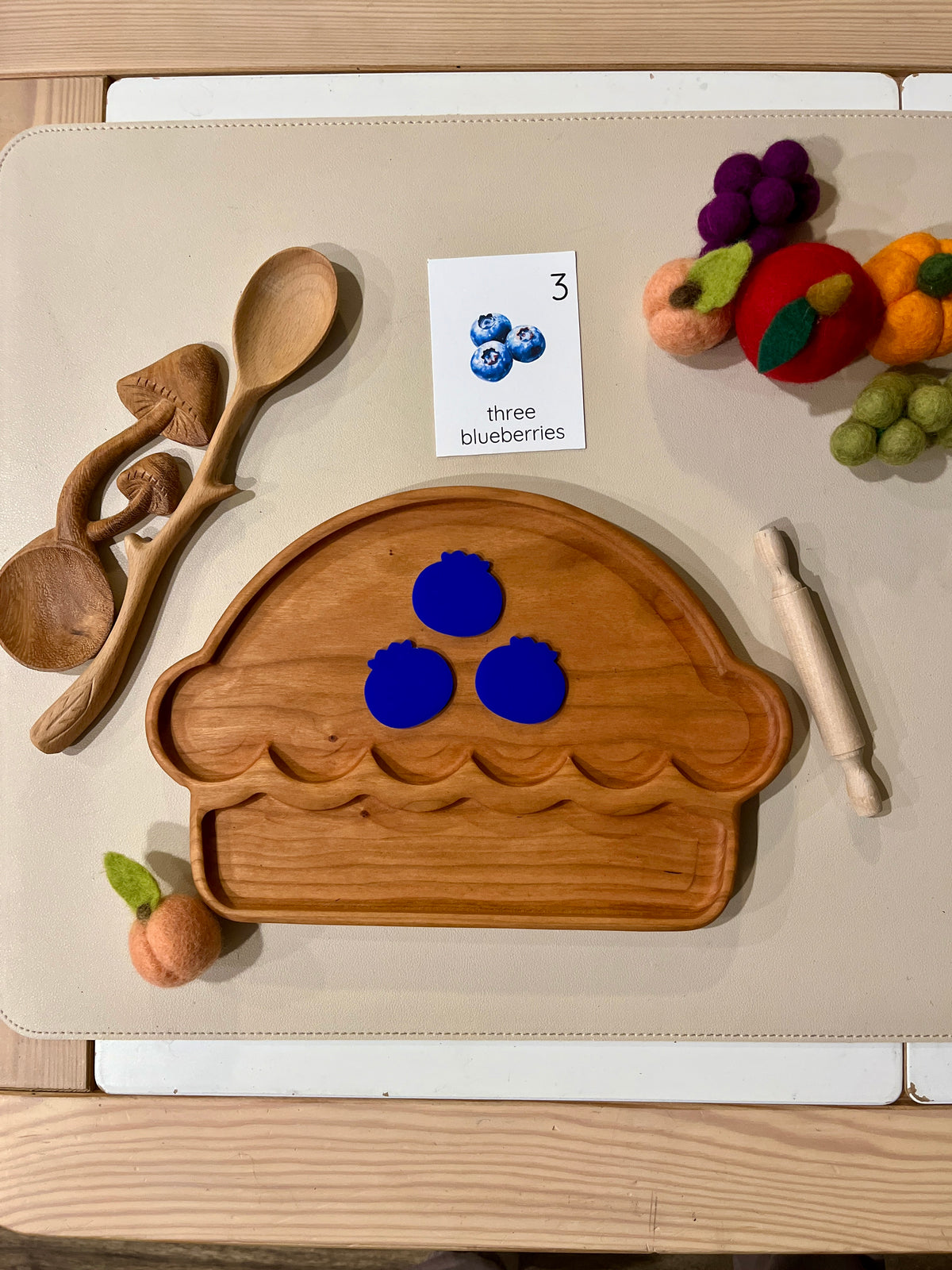 Wooden basket-shaped tray with three blue pomegranate cutouts, wooden spoons, rolling pin, and felt fruit pieces on light surface
