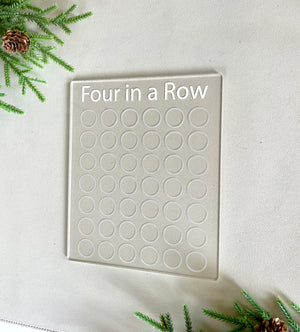 Clear acrylic Four in a Row game board with engraved title and circular slots on a light surface with greenery accents