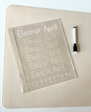 Clear acrylic handwriting practice board with dotted name and numbers alongside a black and white dry erase marker on beige surface