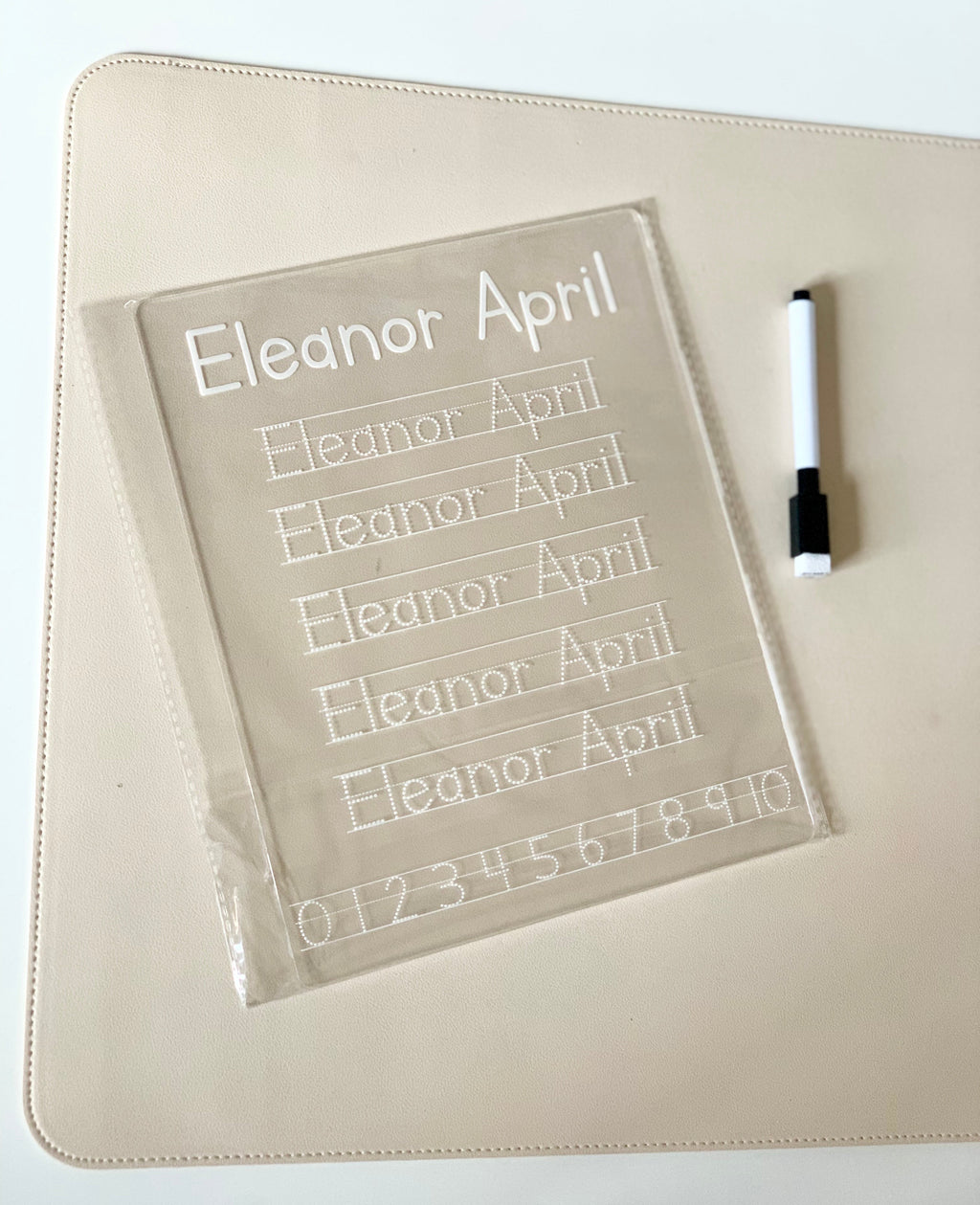 Clear acrylic handwriting practice board with dotted name and numbers alongside a black and white dry erase marker on beige surface