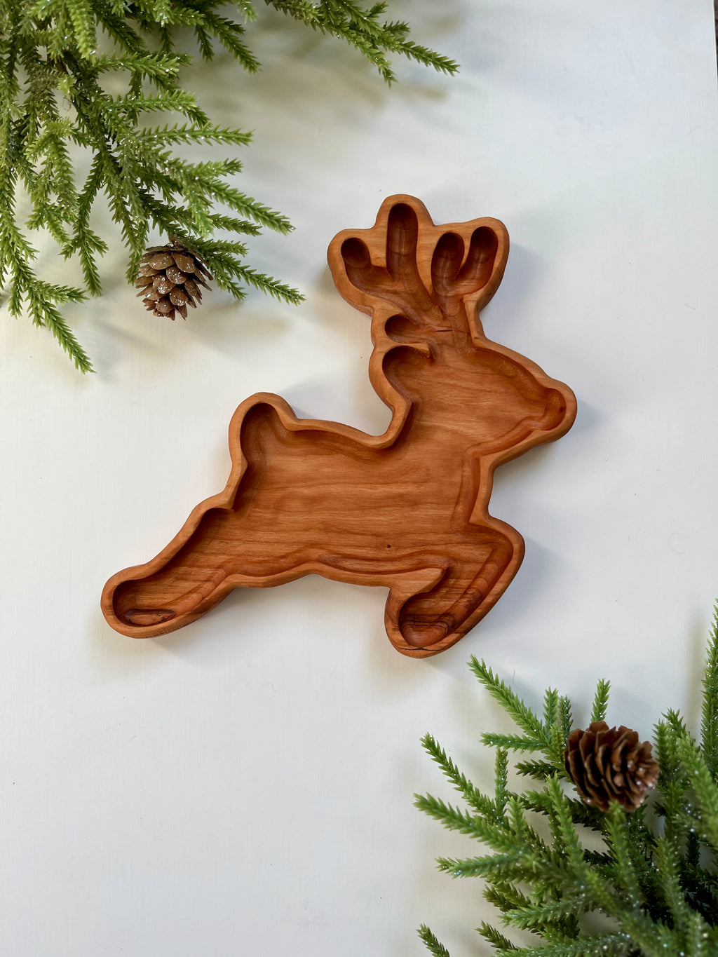 Wooden tray carved in the shape of a reindeer surrounded by green pine branches and pine cones on white surface