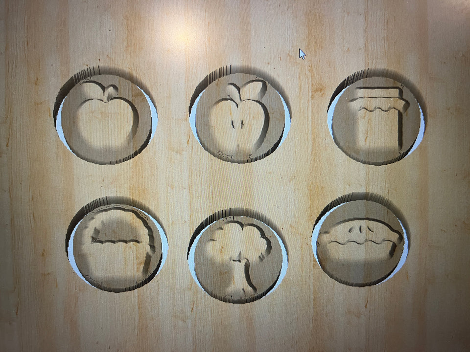 Wooden board with six carved shapes including apple, peach, jam jar, pie slice, tree, and pie on light wood surface