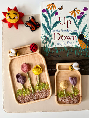 Two wooden trays shaped like jars holding fabric flowers, felt bees, and a ladybug beside a children's book and felt sun and butterfly toys