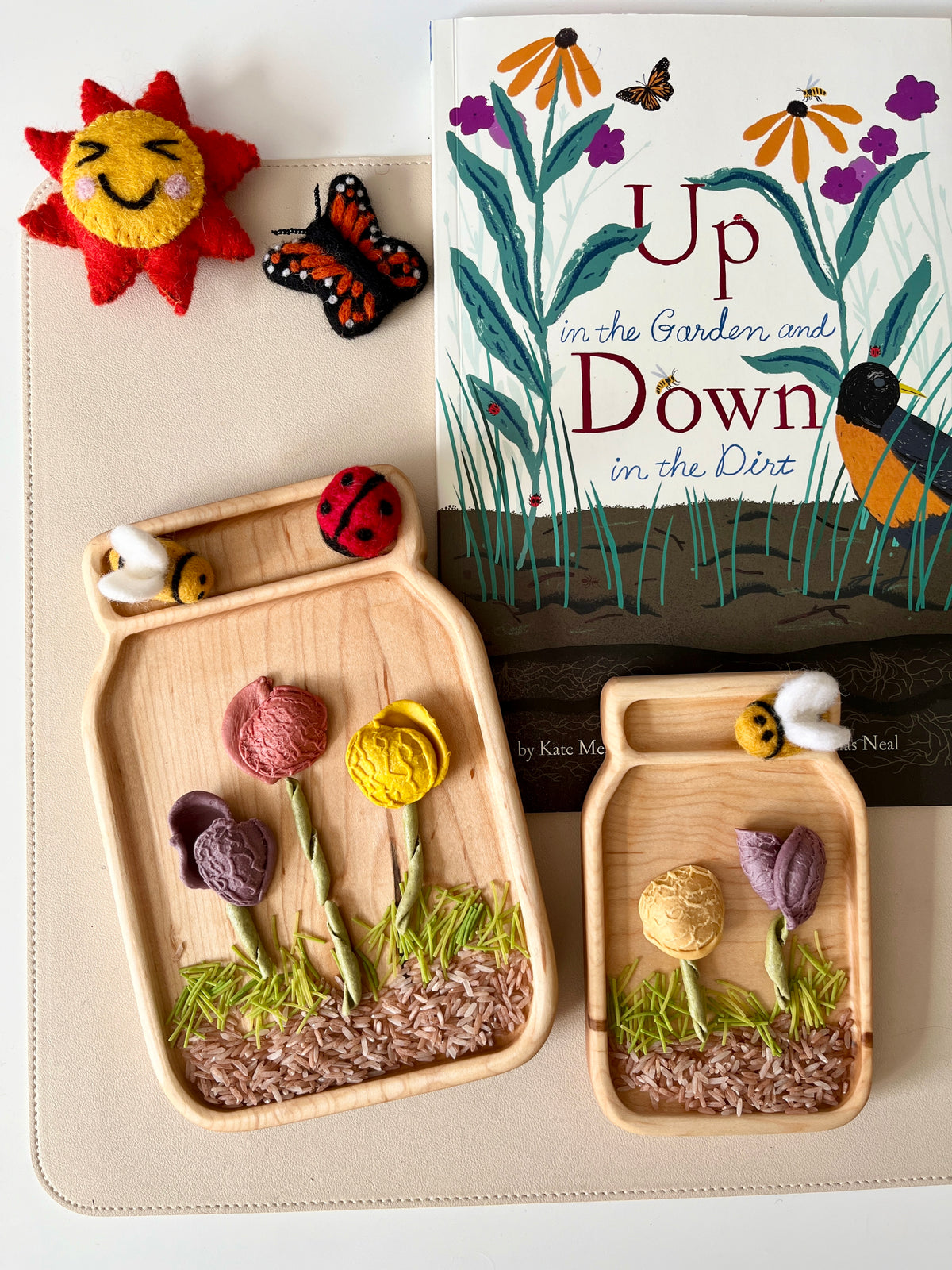 Two wooden trays shaped like jars holding fabric flowers, felt bees, and a ladybug beside a children's book and felt sun and butterfly toys