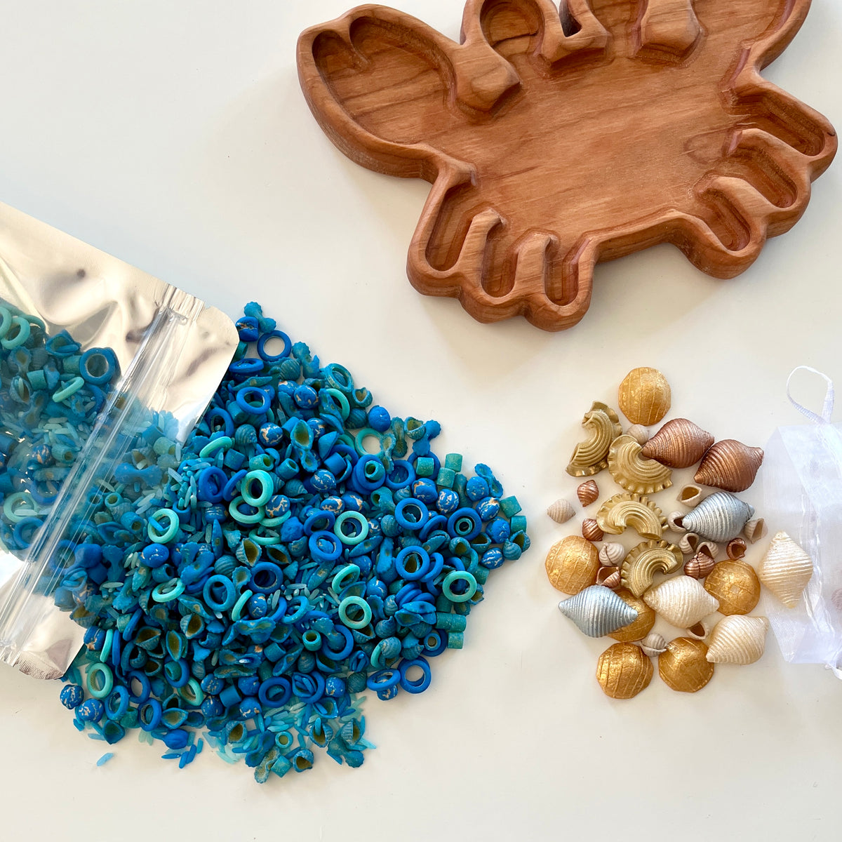 Small blue craft rings spilled from a foil bag near gold, silver, and bronze seashell-shaped beads and a wooden crab-shaped tray