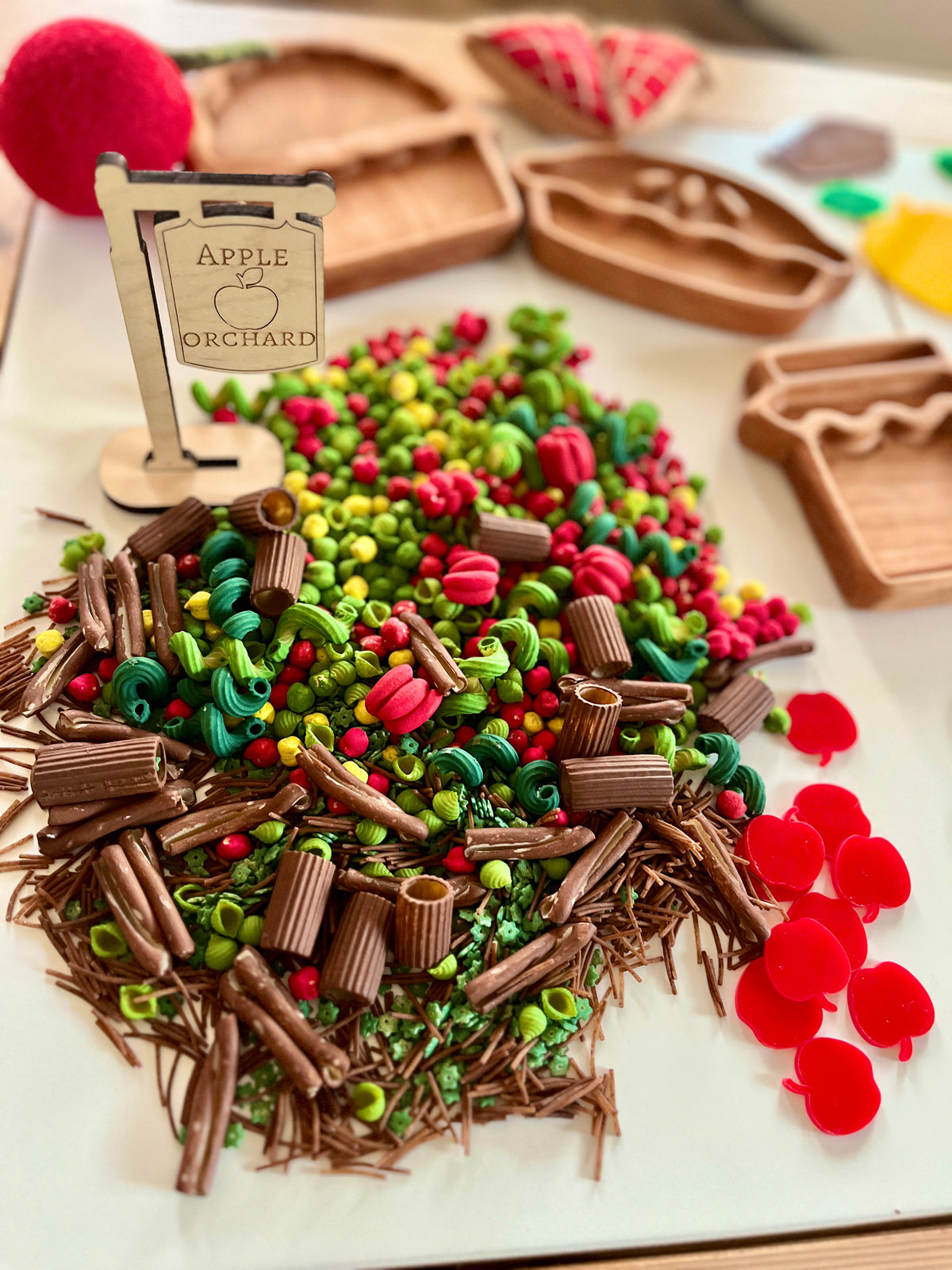 Assorted miniature clay vegetables and twigs with red felt apples and wooden trays on white surface