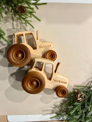 Two handcrafted wooden tractors with personalized names engraved surrounded by pine branches and pinecones on a beige surface