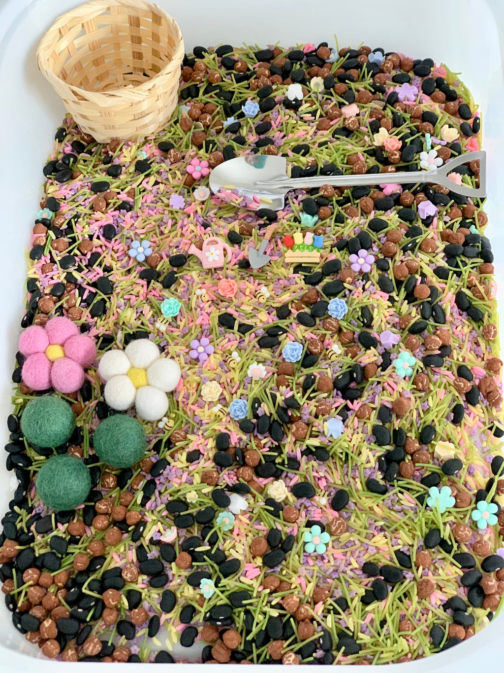 Colorful sensory bin with mixed beans, green pasta, flower-shaped beads, felt balls, small gardening tools, and a wicker basket