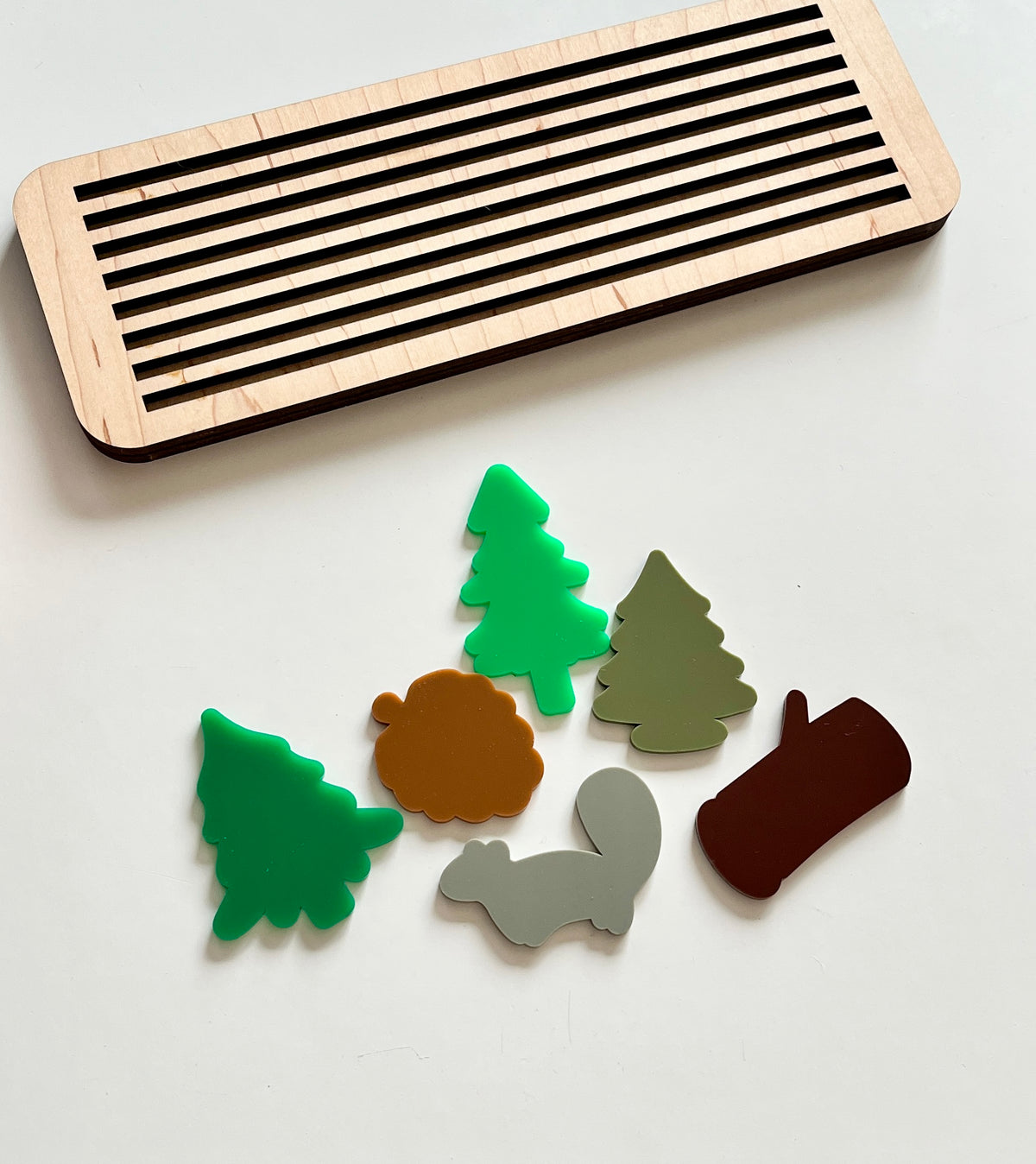 Rectangular wooden tray with black grooves beside colorful acrylic forest shapes including trees, a log, a squirrel, and a pinecone