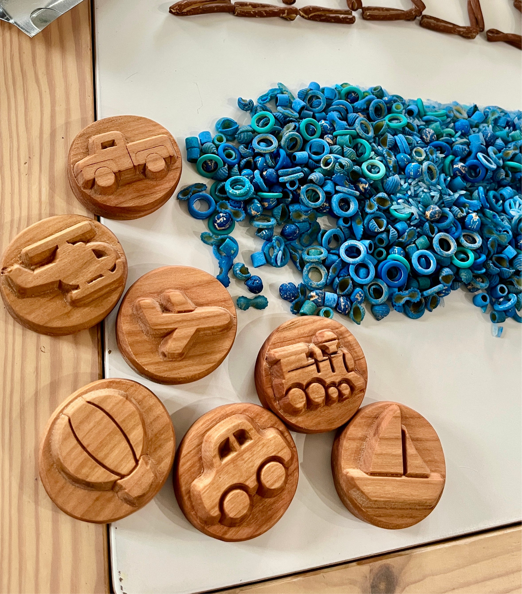 Round wooden toys carved with transportation shapes including car, airplane, boat, and helicopter beside blue textured beads on table