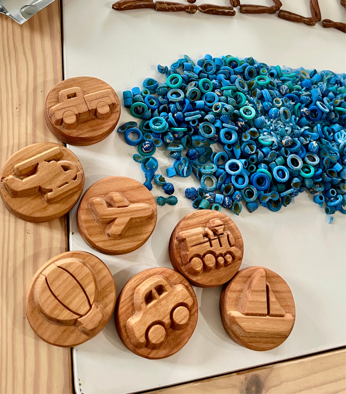 Round wooden toys carved with transportation shapes including car, airplane, boat, and helicopter beside blue textured beads on table