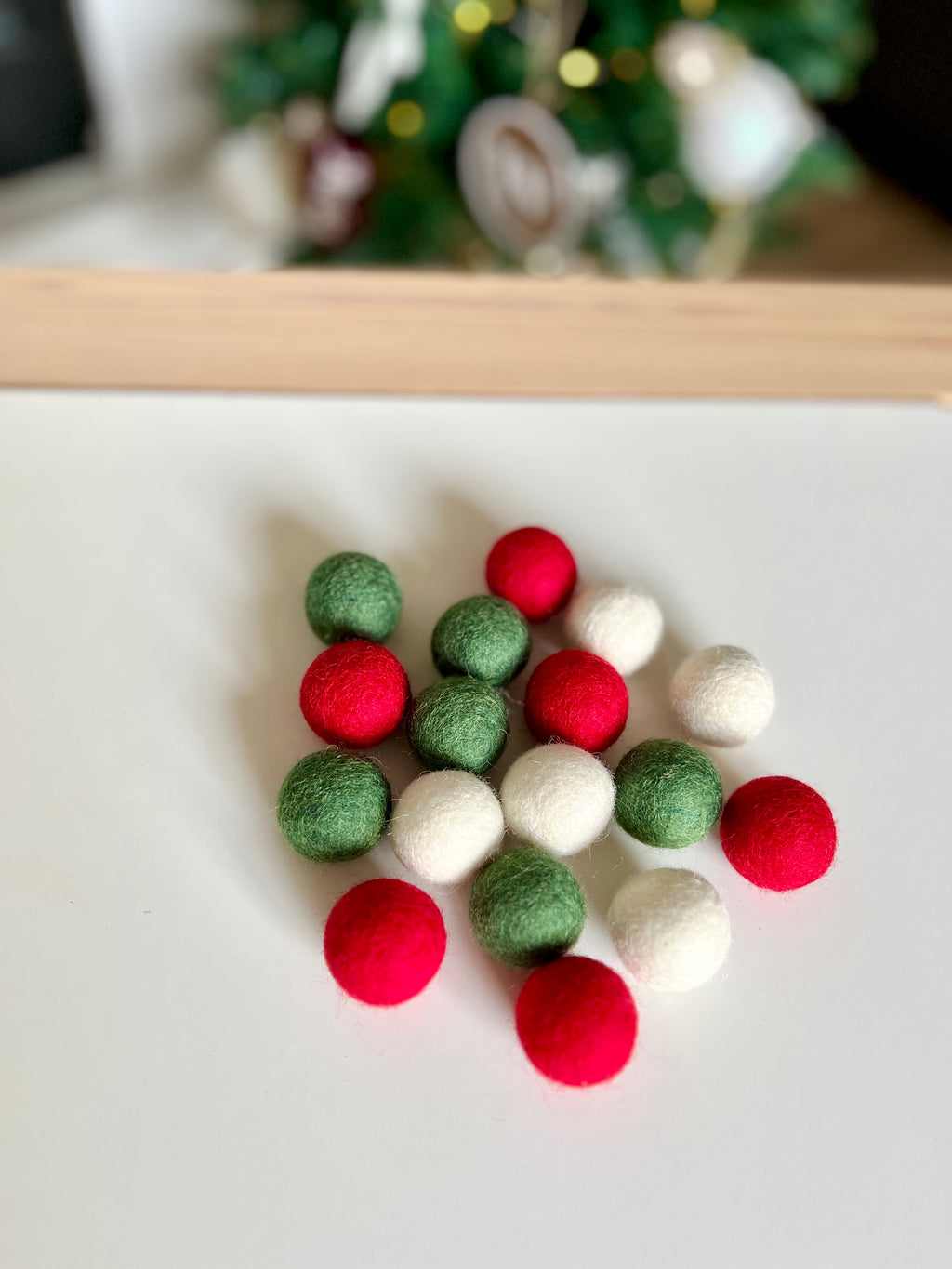 Small red, green, and white felt balls arranged on a white surface with blurred holiday decorations in the background