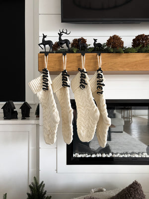 Four cream cable knit Christmas stockings with black cursive name tags hanging from wooden mantel decorated with black deer and greenery
