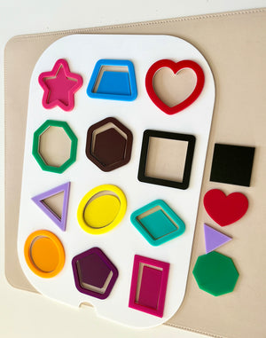 Colorful geometric plastic shapes arranged on a white board with additional shapes on a beige surface nearby