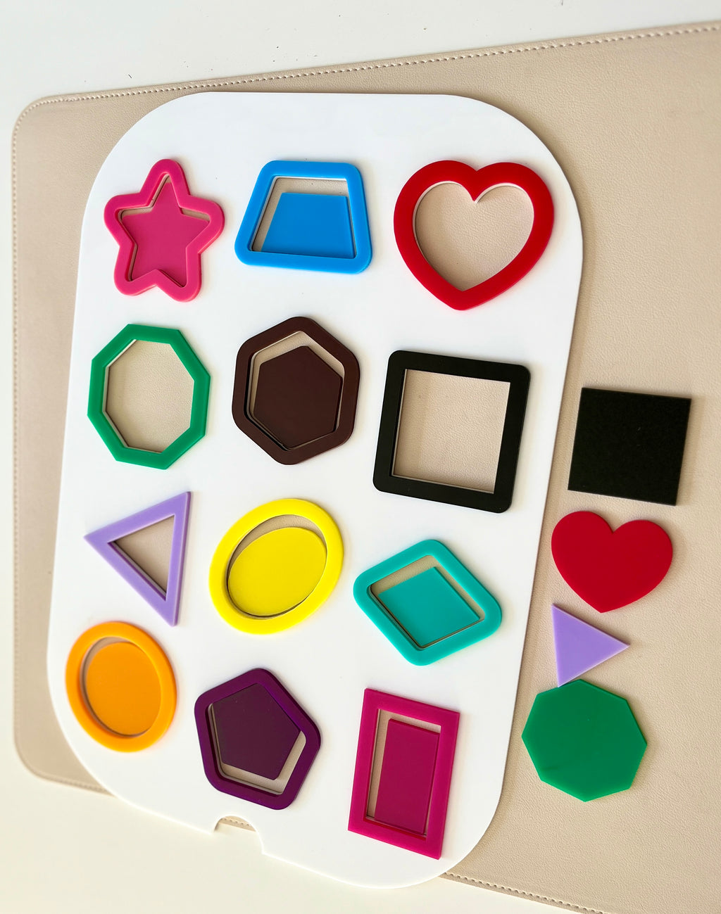 Colorful geometric plastic shapes arranged on a white board with additional shapes on a beige surface nearby