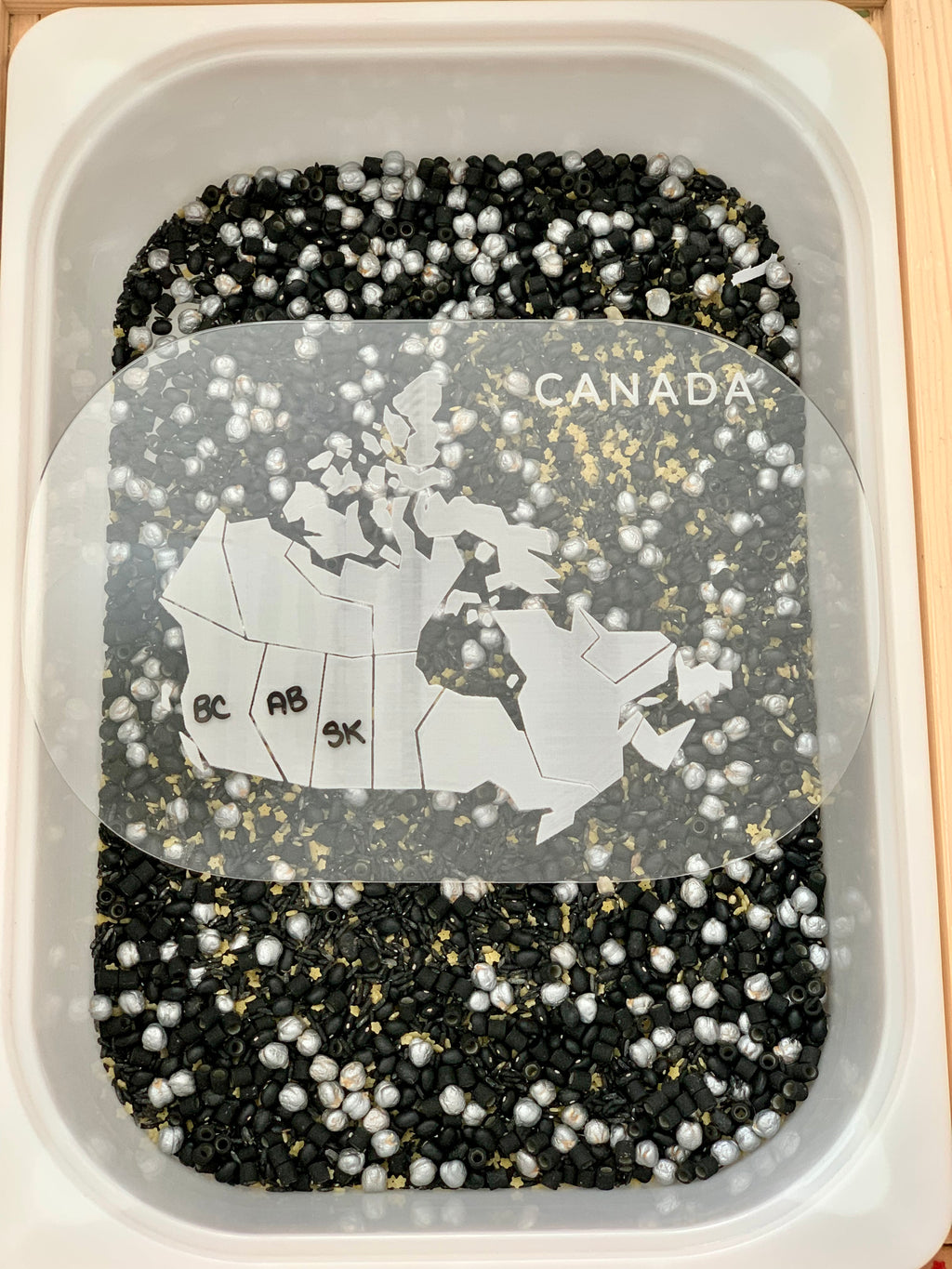 Sensory bin filled with black and white beads featuring a clear map of Canada with labeled provinces inside the container
