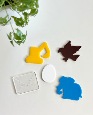 Colorful acrylic shapes including yellow excavator, brown bird, clear envelope, white egg, and blue elephant on white surface with green plant nearby