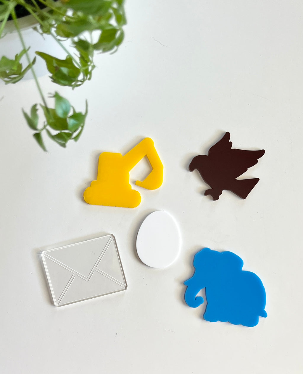 Colorful acrylic shapes including yellow excavator, brown bird, clear envelope, white egg, and blue elephant on white surface with green plant nearby