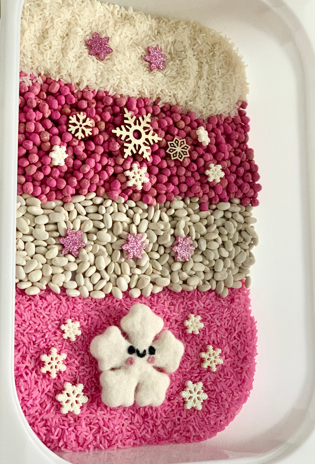 Winter sensory tray with pink rice, white beans, pink peas, wooden snowflakes, and felt snowflake figure on white tray