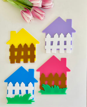 Four colorful house-shaped cutouts with white or brown fences and green grass pieces beside pink tulips on beige surface