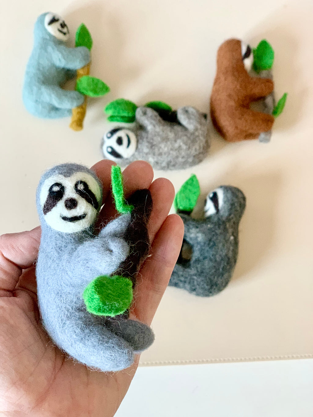 Hand holding a small gray felt sloth figurine clinging to a branch with green leaves with four other felt sloths in the background