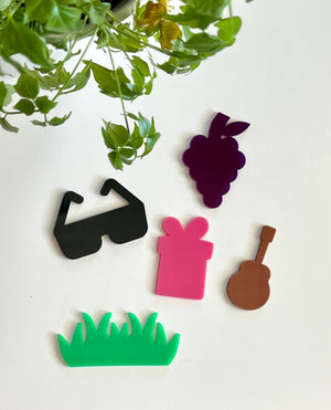 Five colorful acrylic shapes including black sunglasses, purple grapes, pink gift box, brown guitar, and green grass under hanging green leaves