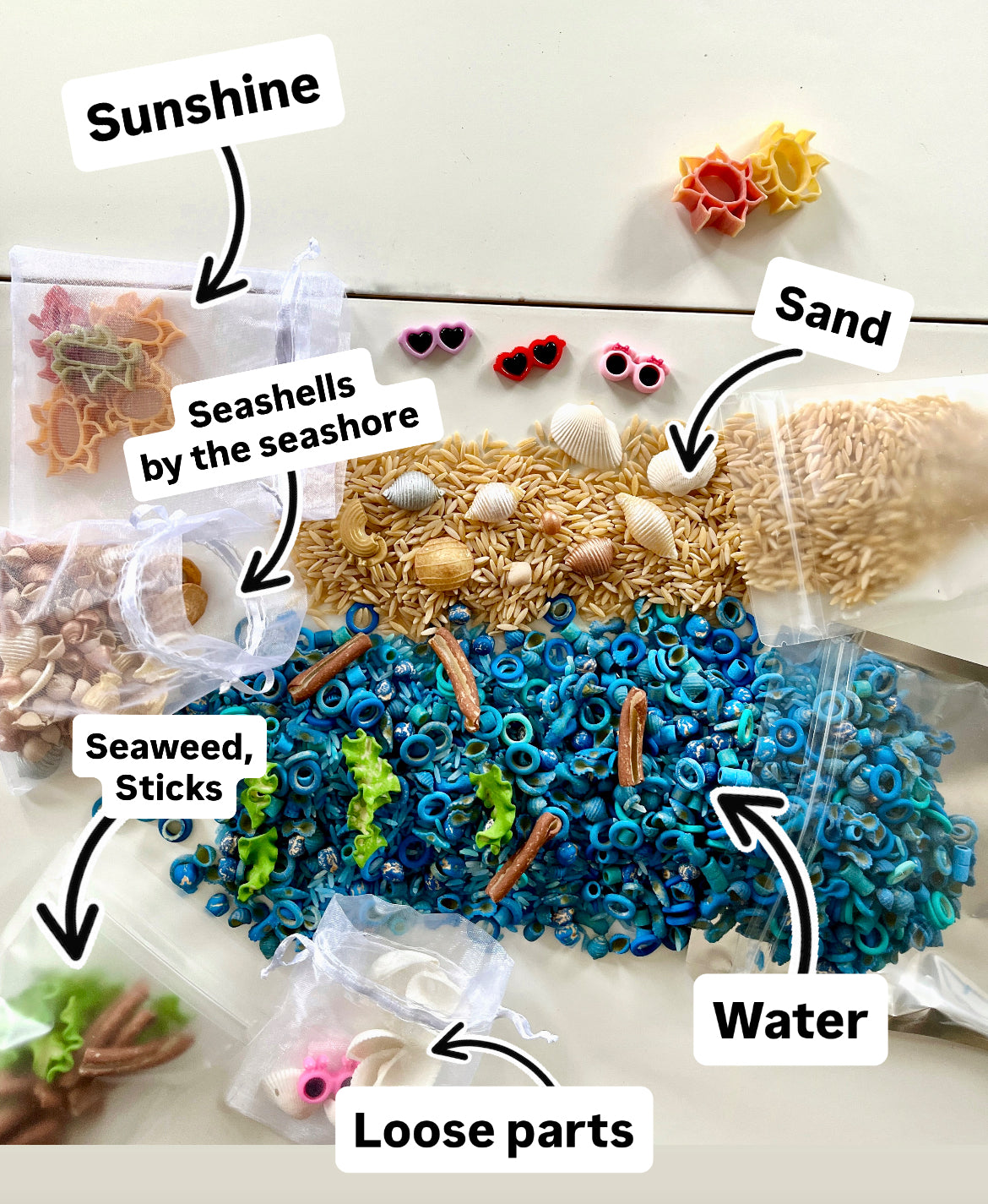 Assorted small shells, beads, pasta, and sticks arranged to resemble a beach scene with labeled clear pouches