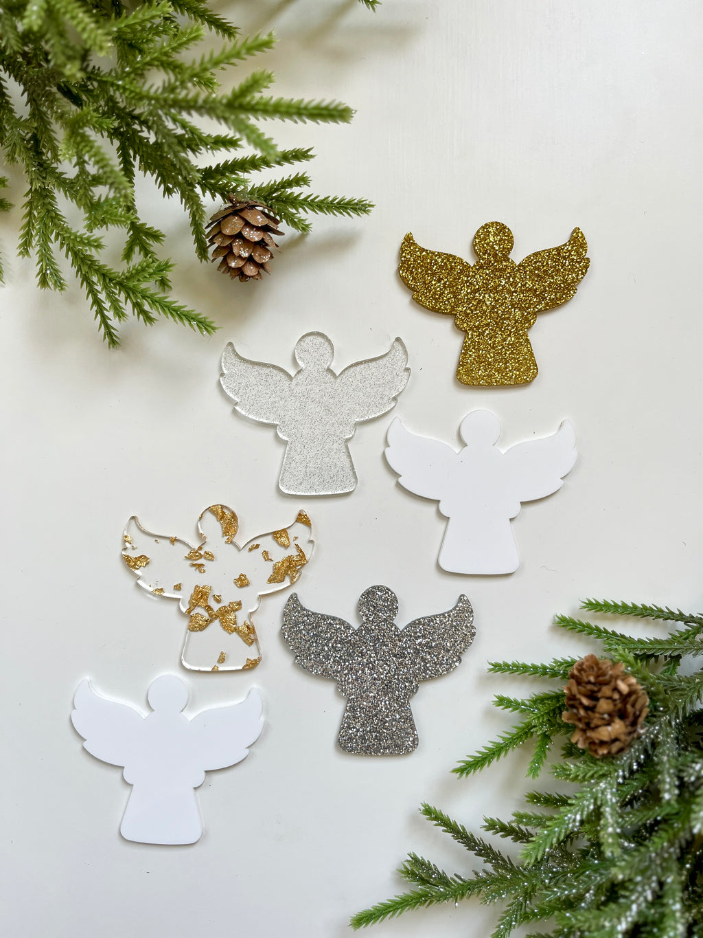 Six angel-shaped cutouts in white, gold, silver glitter, and gold foil on white background with pine branches and pinecones