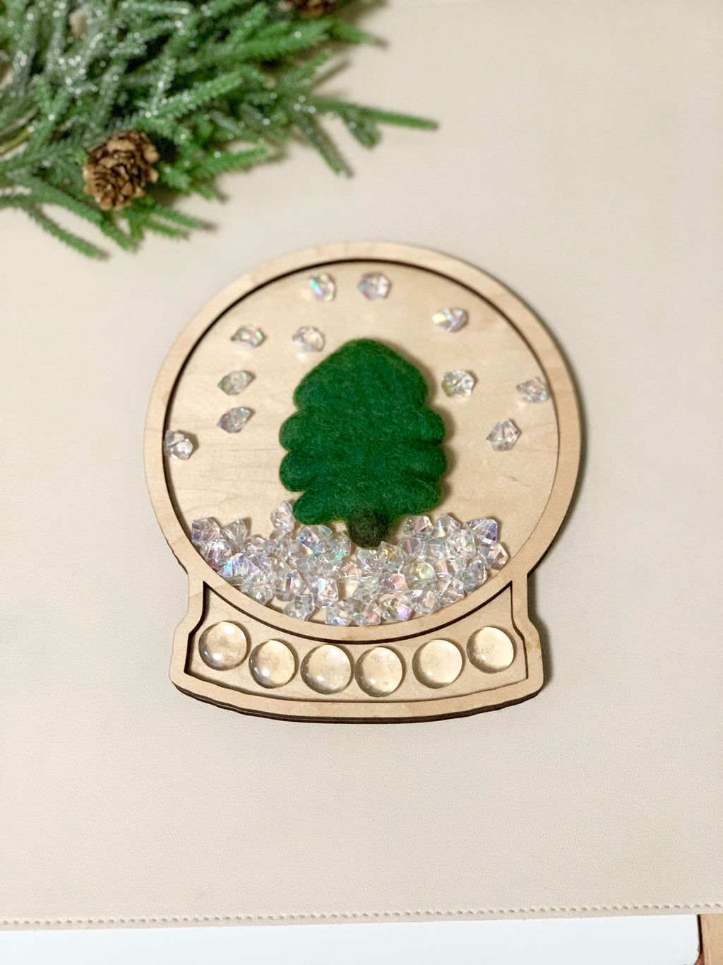 Wooden snow globe-shaped sensory tray with green felt tree and clear acrylic gems on light surface with pine branch