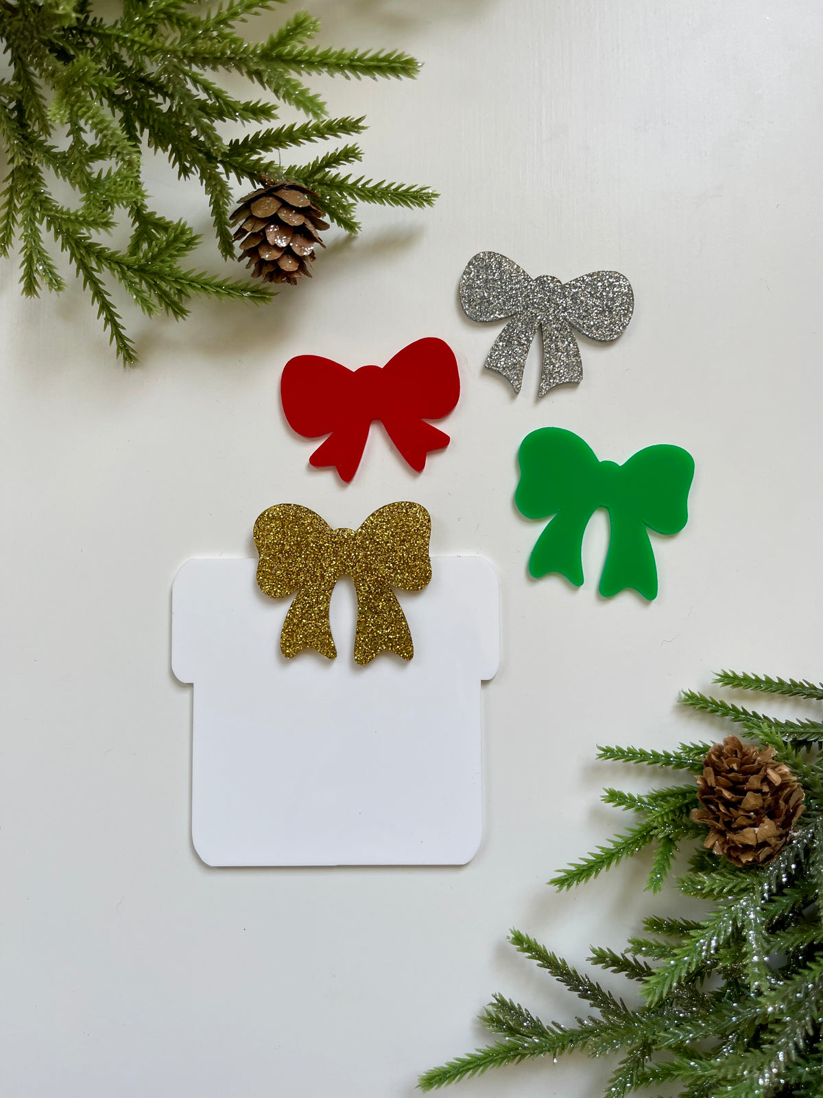 Four decorative acrylic bows in red, green, silver glitter, and gold glitter on white background with pinecones and greenery