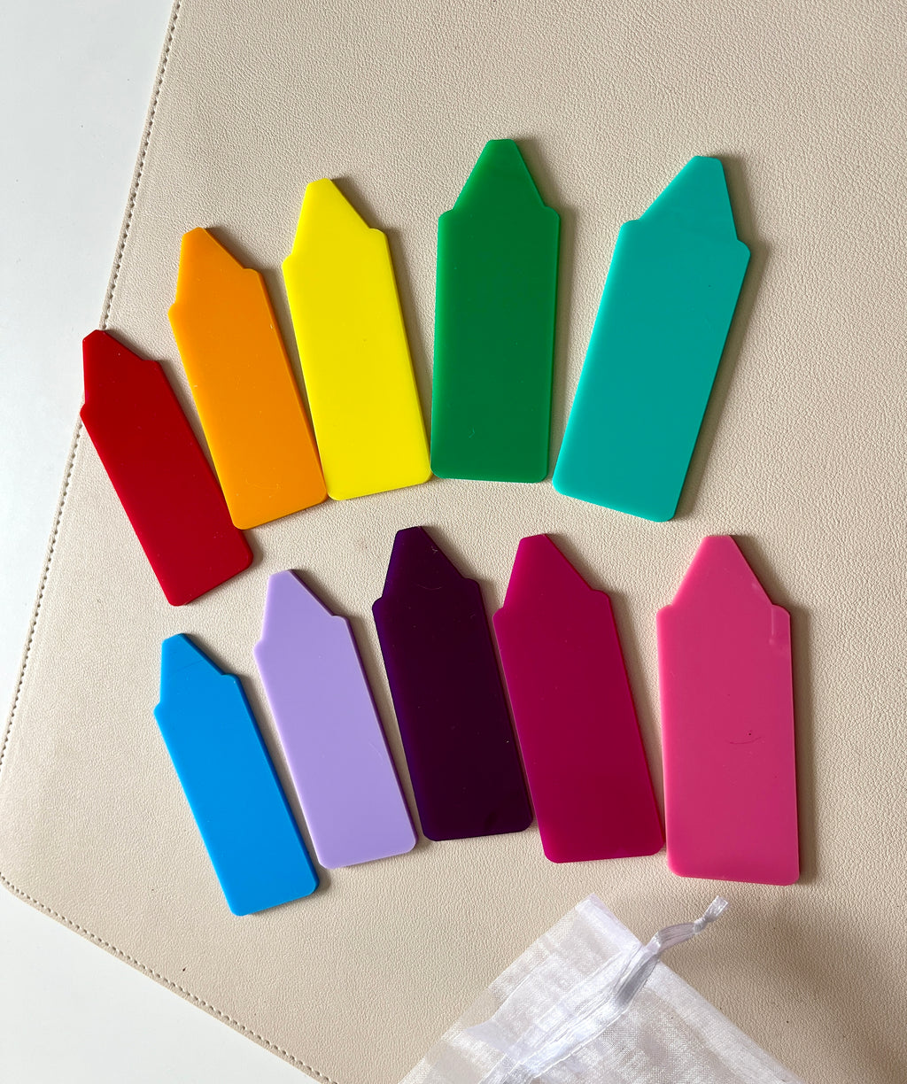 Plastic plant labels in a rainbow of colors arranged on a beige surface alongside a white drawstring bag