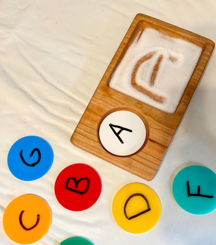 A wooden salt and sand tray with a slot for letters or numbers, placed on a surface with colorful circular markers corresponding to the alphabet.
