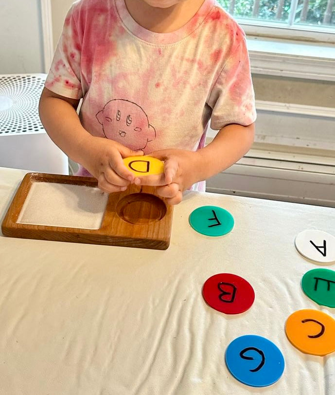Child wearing a pink tie-dye shirt holding yellow letter disc over wooden tray with colorful alphabet discs on table