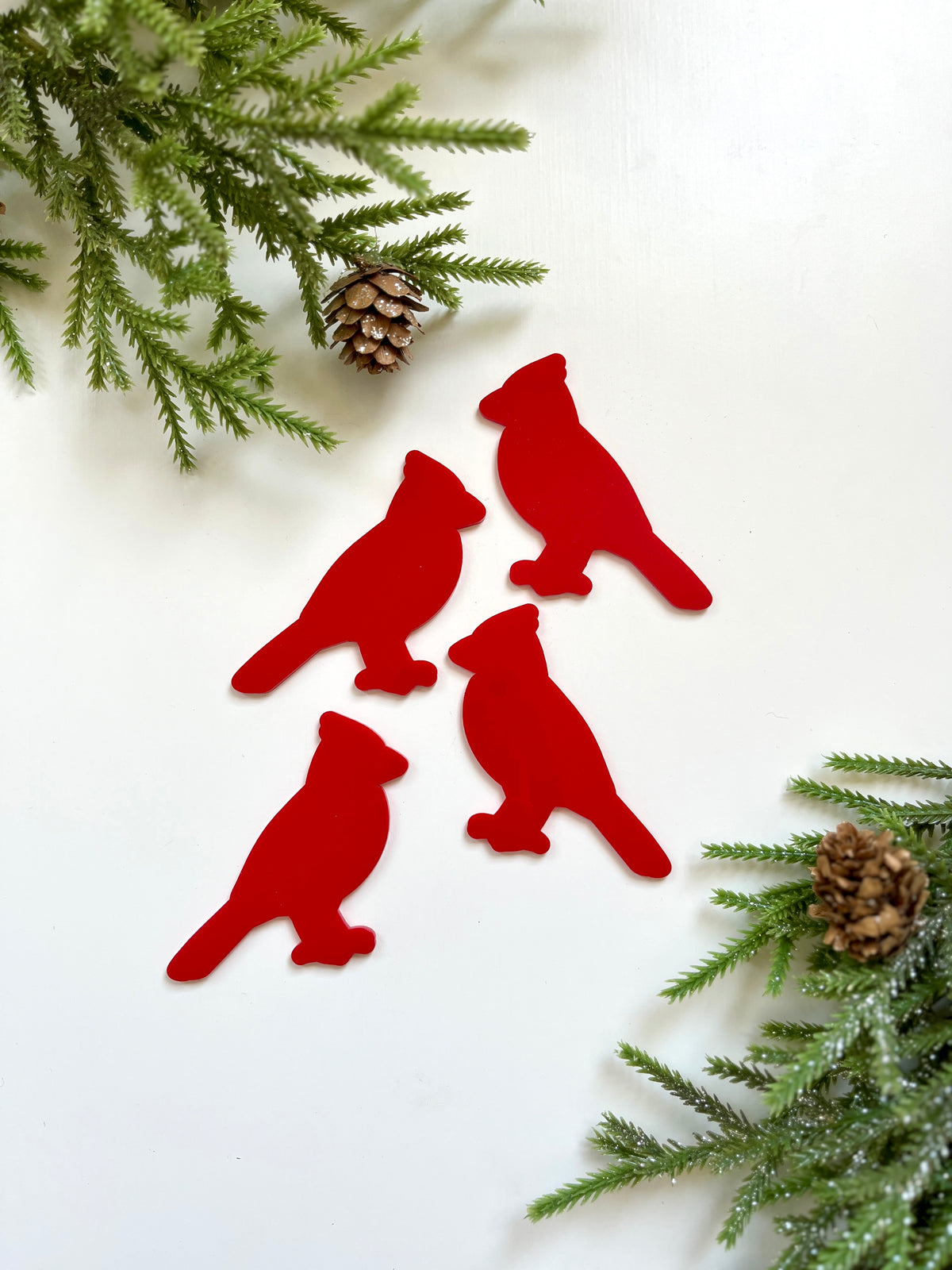 Four red cardinal bird-shaped cutouts surrounded by frosted pine branches and pinecones on white background