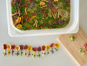 Colorful handmade felt petals and leaves arranged on green moss inside a white container and wooden board on a white surface