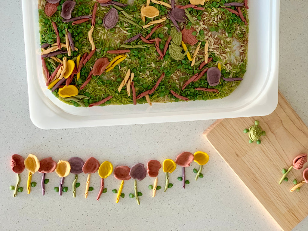 Colorful handmade felt petals and leaves arranged on green moss inside a white container and wooden board on a white surface