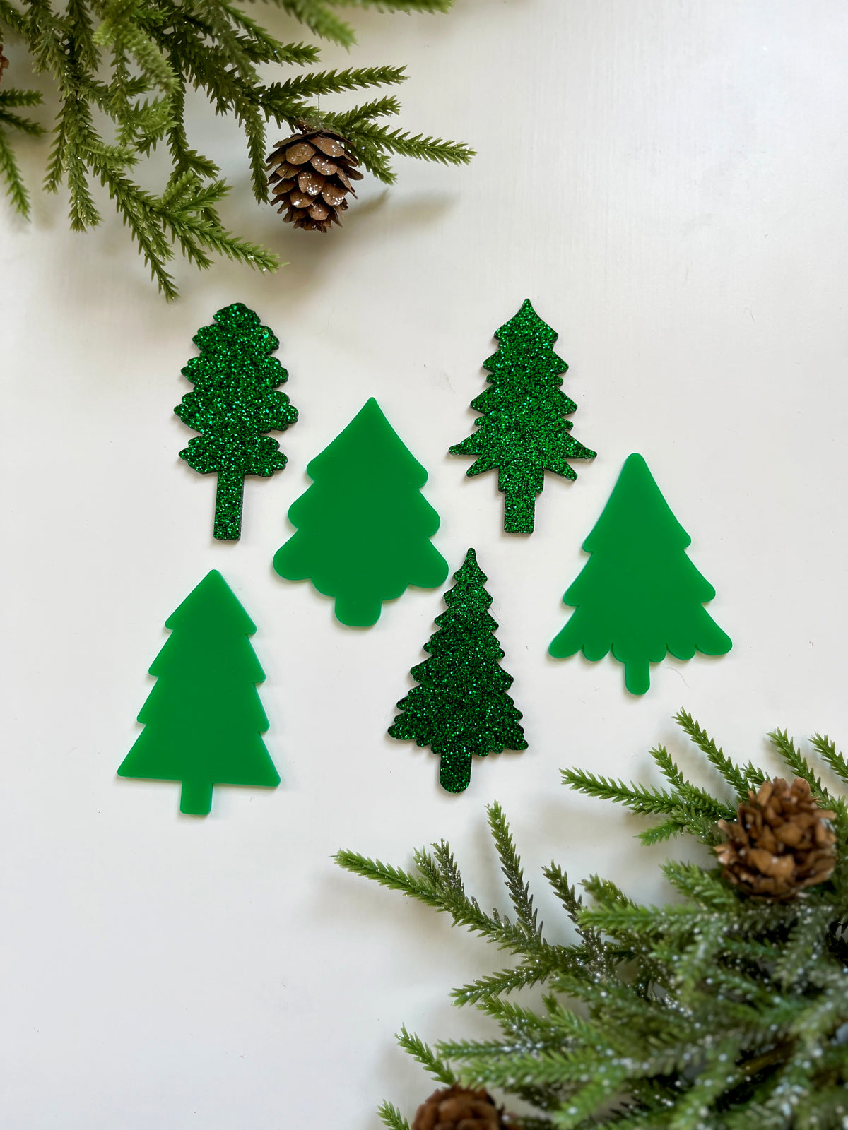 Six assorted green Christmas tree-shaped cutouts with matte and glitter finishes surrounded by pine branches and pinecones on white background