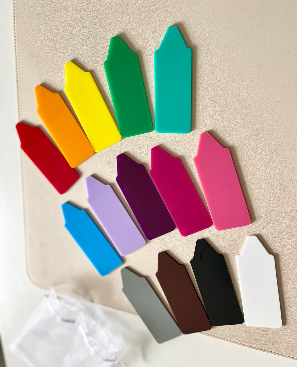 Multi-colored plastic plant markers arranged in a loose arc on a light beige surface with a white drawstring bag nearby