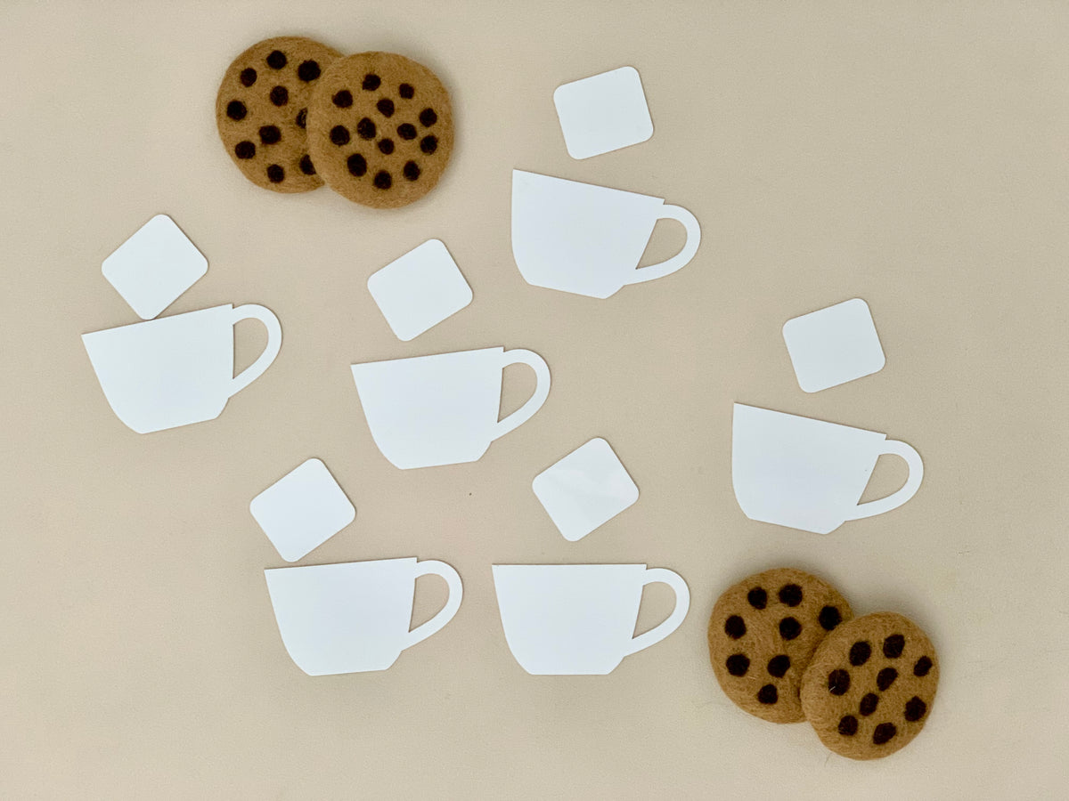 White paper cutouts of coffee cups and sugar cubes arranged with felt chocolate chip cookie shapes on beige background
