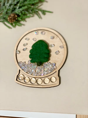 Laser cut wooden snow globe sensory tray with felt tree, clear gems, and pine branch on neutral background